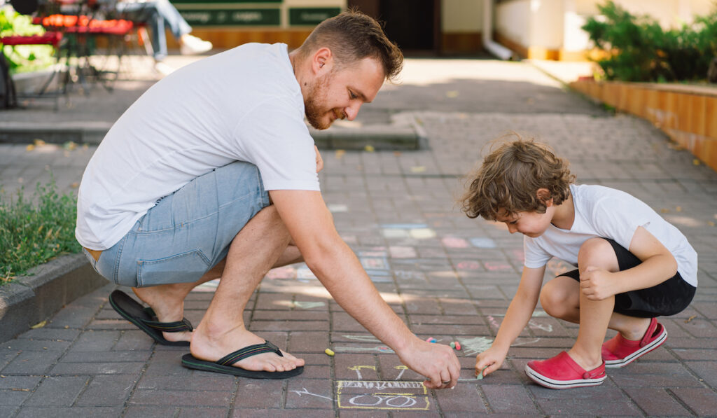 A little preschool boy with father draws with colorful chalks on the ground
