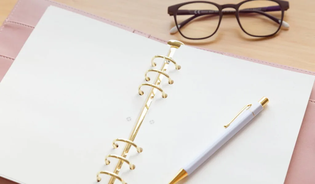 An open pastel pink notebook with pen and glasses on a wooden desk. 