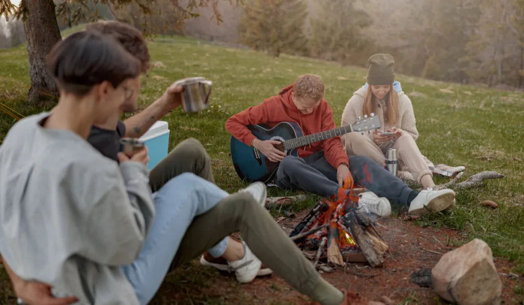 Cheerful friends hanging out together at nature and fire with guitar playing and singing.
