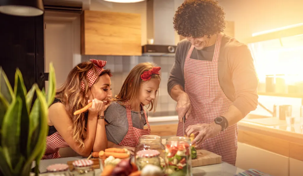 Cute little girl and her beautiful parents are cutting vegetables and smiling while making pickle in kitchen at home

