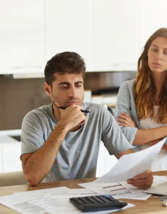 Frustrated young European couple facing financial crisis. Man with stubble sitting at kitchen table and studying notification from bank, his stressed wife standing behind him with arms crossed