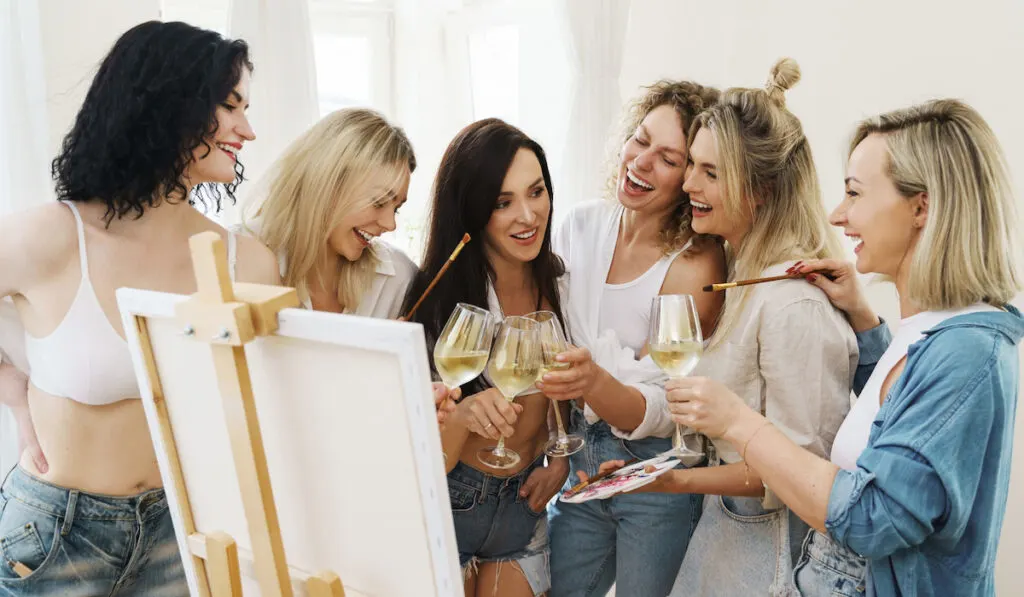Group of young beautiful women paint on canvas and drinking white wine during party at home
