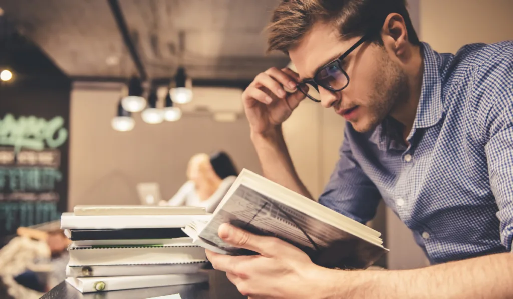 Concentrated young man is reading a book while working hard in the modern library

