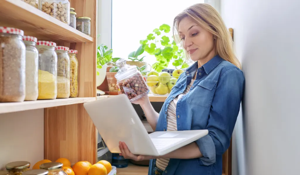 woman holding a jar of peanuts and checking her laptop for ingredients