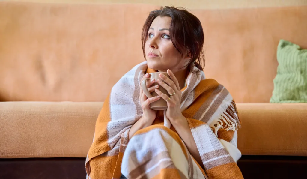 Middle-aged woman under warm woolen blanket with cup of hot drink
