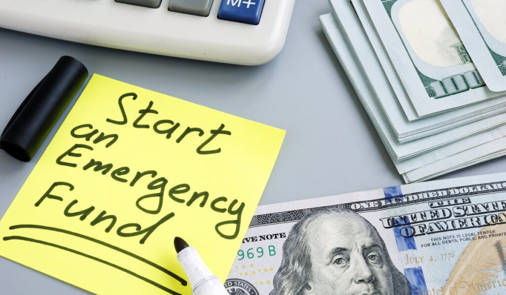 start an emergency fund written on a sticky notepad, with dollars in cash beside it 