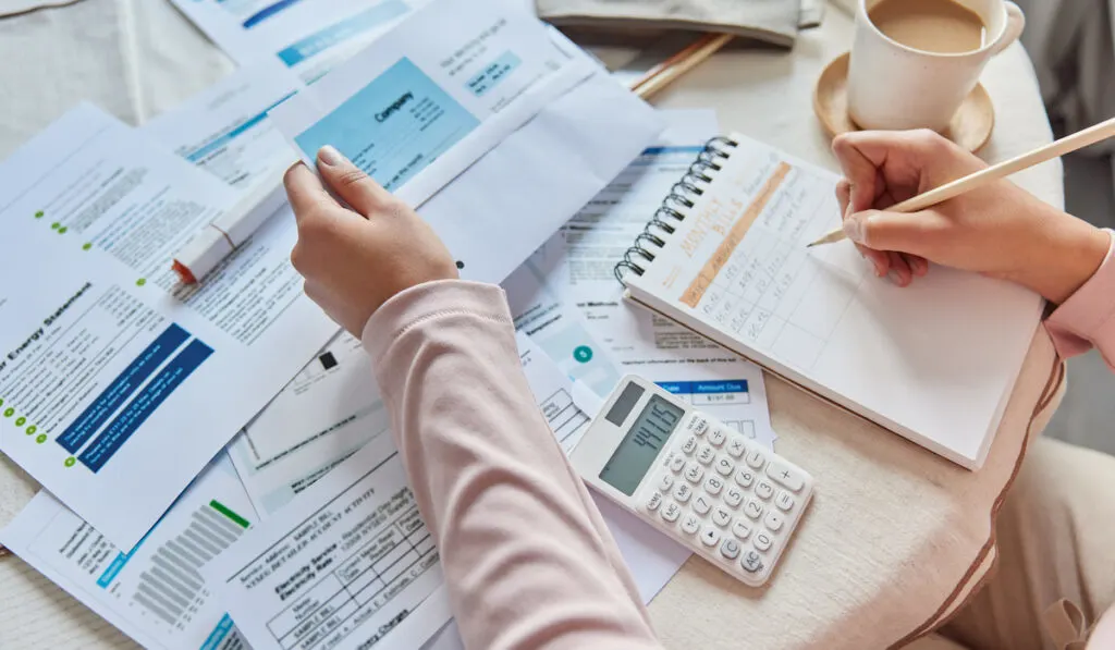 Unrecognizable woman writes down monthly bills in notebook with calculator and coffee on the table