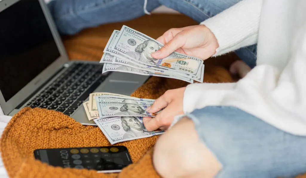 Woman counting her money on her bed with cellphone as her calculator and her laptop