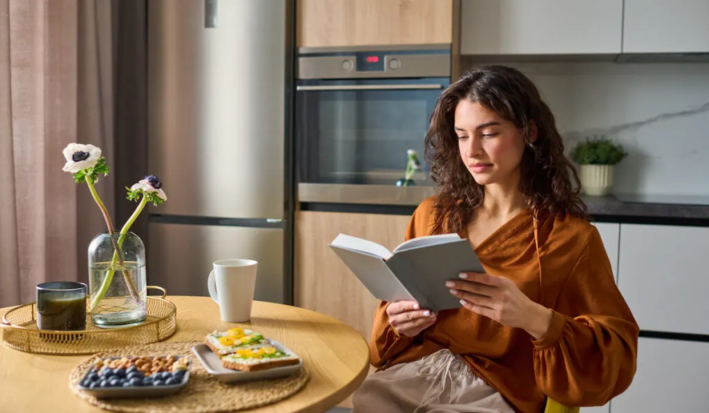 Young relaxed woman in casualwear reading book while sitting by kitchen table