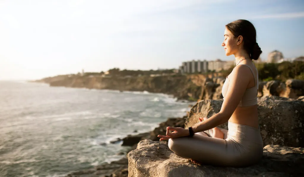 Young woman in sportswear meditating in front of sunrise on beach shore