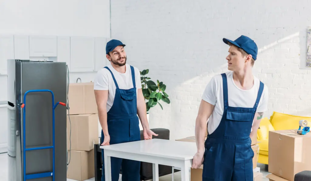 two movers in uniform transporting table in apartment
